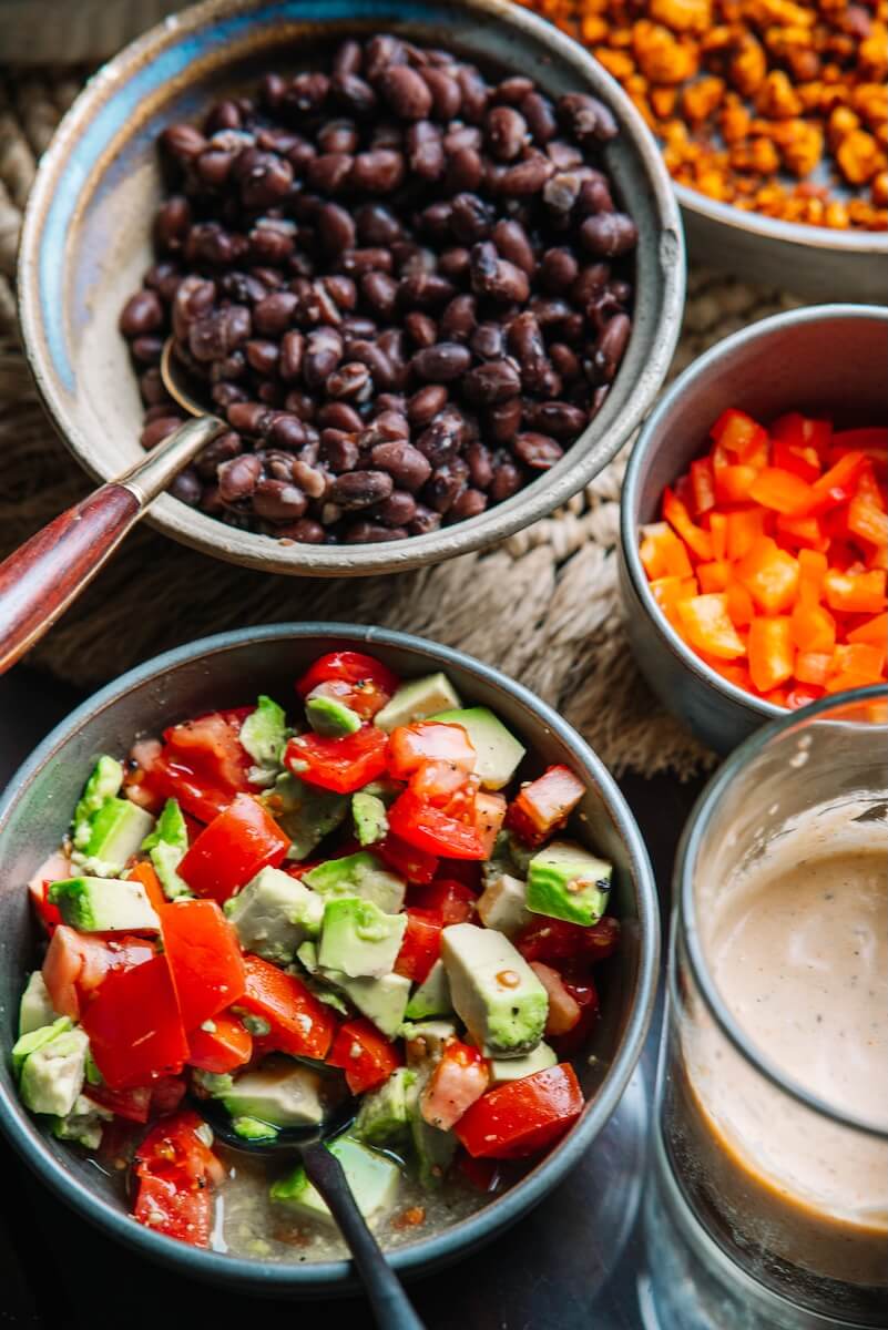 RECIPE: Vegan Taco Bowl - 5 A colorful Vegan Taco Bowl features black beans, diced tomatoes, and avocado, vibrant orange bell peppers, and roasted chickpeas. In the foreground gleams a glass of dressing ready to drizzle over this delightful vegan creation.