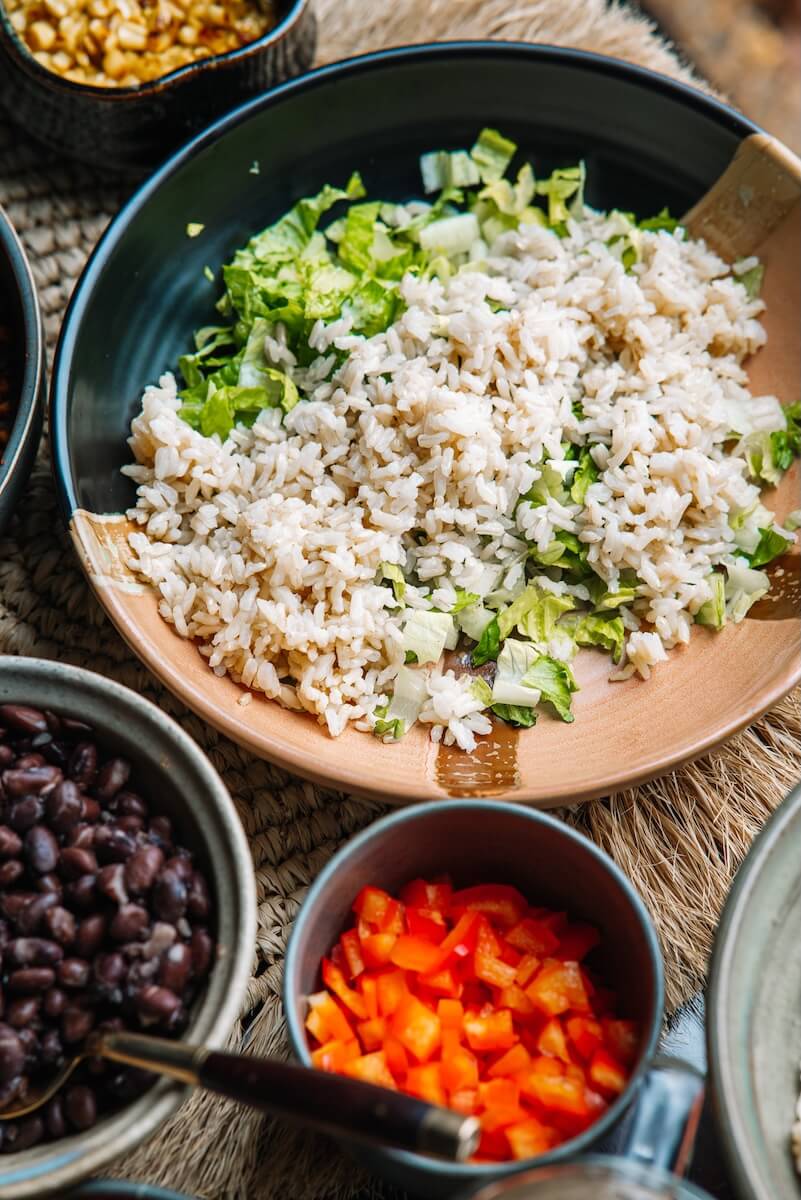 RECIPE: Vegan Taco Bowl - 3 A vibrant vegan bowl featuring rice and chopped lettuce, surrounded by bowls of black beans, diced red bell peppers, and other colorful ingredients that bring a taco-inspired flair to your meal.