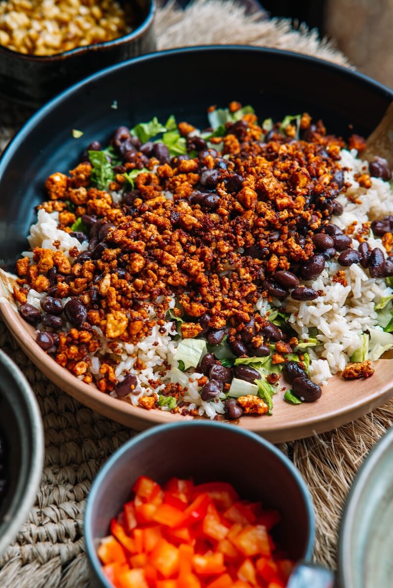 RECIPE: Vegan Taco Bowl - 4 A vibrant vegan bowl featuring spicy crumbled tofu, black beans, and cilantro over a bed of rice, accompanied by a side of chopped red bell peppers.