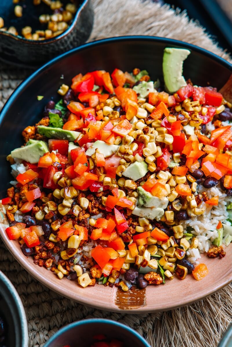 RECIPE: Vegan Taco Bowl - 6 A vibrant taco bowl featuring a base of fluffy rice, layered with black beans, diced red peppers, grilled corn, creamy avocado slices, and crumbled cheese.