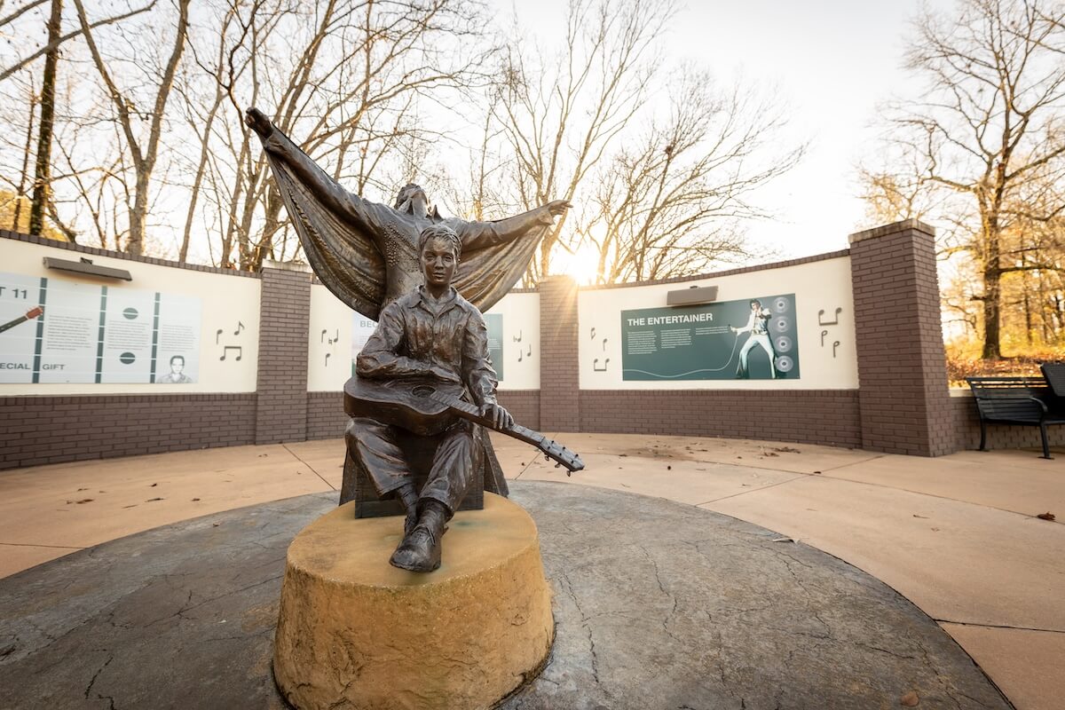 In the birthplace of American music, a bronze statue of a person sits with a guitar, embraced by a wall adorned with musical information panels, all nestled in a tranquil park setting.