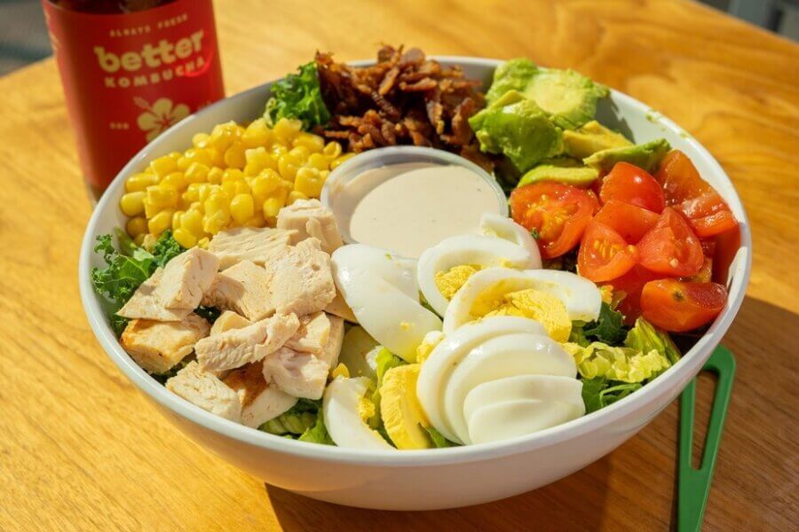 The Best Salads in BHAM: 16 Spots We Love - 5 A bowl of salad with sliced hard-boiled eggs, cherry tomatoes, avocado, corn, grilled chicken, bacon bits, lettuce, and a container of dressing at the center—just what you'd expect from the best salads in Birmingham. A bottle of kombucha is nearby.