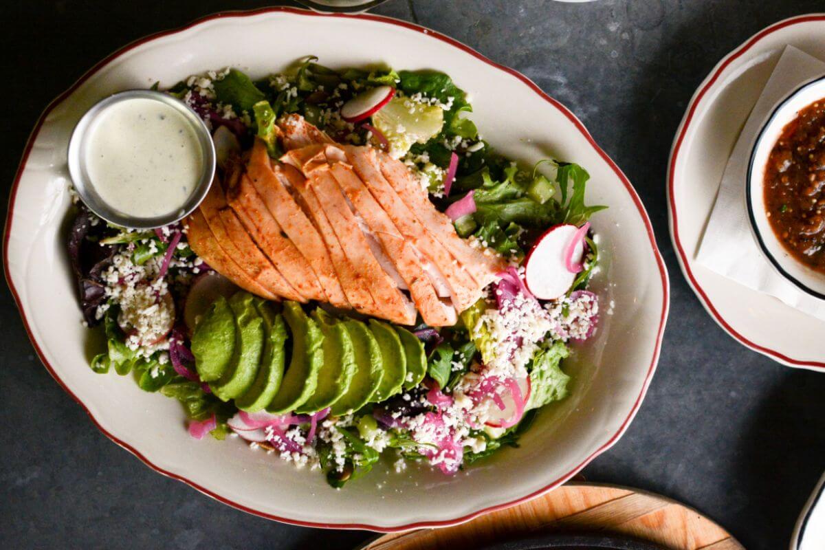 A plate of mixed green salad topped with sliced grilled chicken, avocado, radishes, cheese, and a side of dressing, plus a bowl of salsa—one bite and you'll see why it's among the best salads in Birmingham.