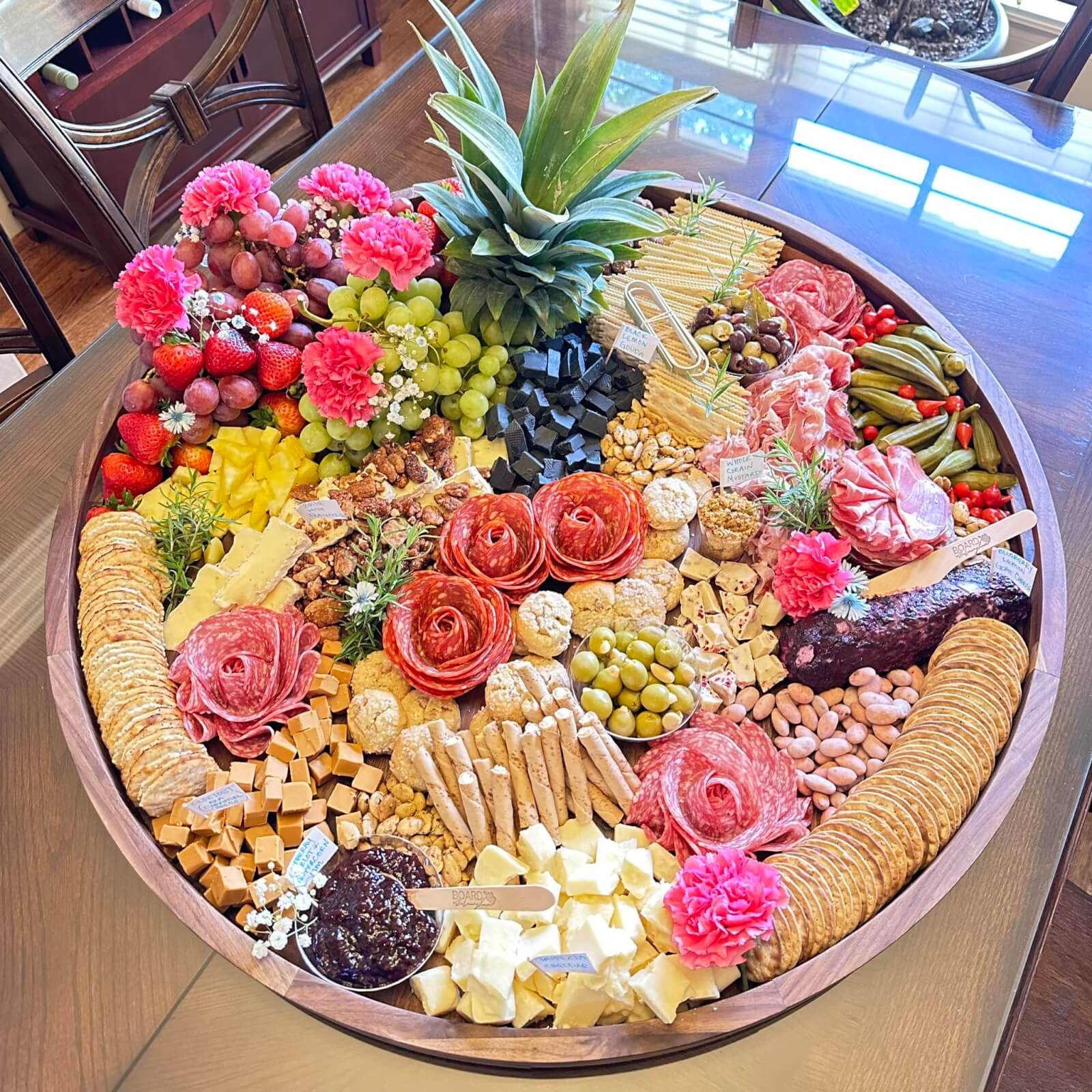 A large round charcuterie board with an assortment of cheeses, meats, crackers, fruits, and garnishes, including olives, grapes, strawberries, and decorative flowers.