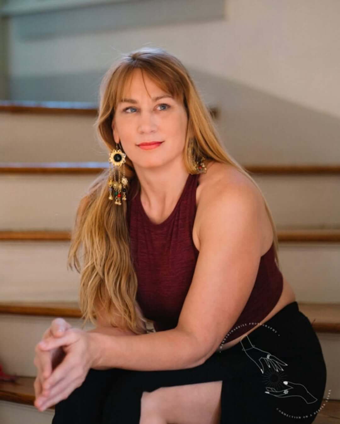 Bridget Herring sits on the stairs at The Oha in a sleeveless maroon top and large earrings, looking ahead with serene focus.