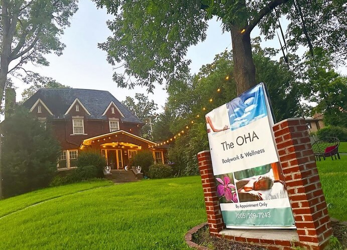 Brick house surrounded by trees, with a sign in the yard reading "The Ōha Bodywork & Wellness." String lights are hung above the entrance, welcoming you to this serene space where skilled massage therapists offer their healing touch.
