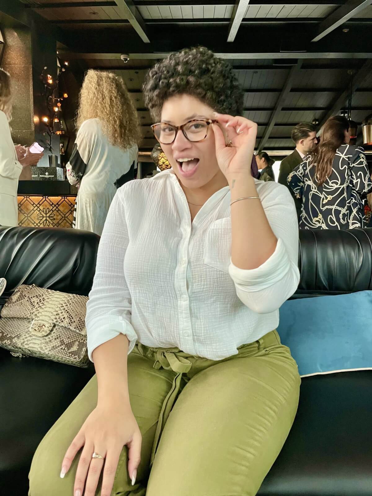 A person in a white shirt and green pants sits on a black couch, playfully sticking their tongue out and holding glasses with one hand. 