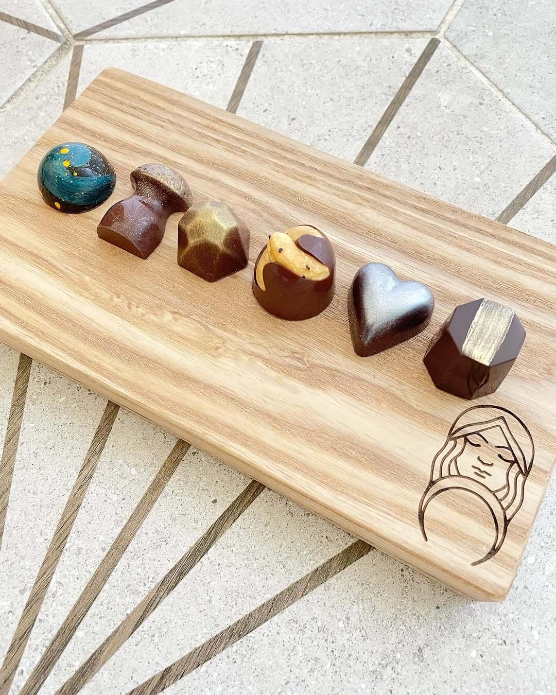 A wooden board artfully showcases six assorted Divine Chocolates from Chocolatá, referred to as a 'Chocolate flight,' each with unique shapes and designs, elegantly set on a geometric patterned surface.