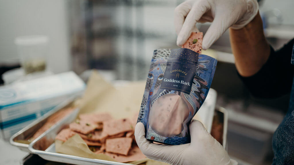 A person in gloves holds a package labeled "Goddess Bark" with one piece being put in, as the same Divine Chocolates rest enticingly on a tray in the background.