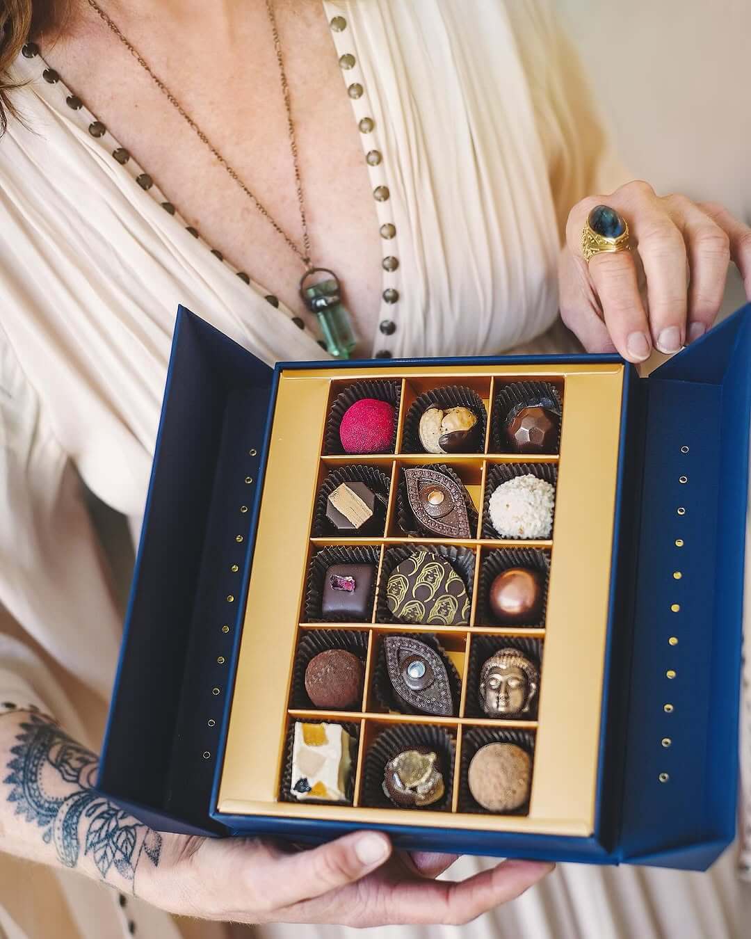 Kathy D'Agostino holds an open box of divine Chocolatá chocolates, showcasing assorted treats with various shapes and elegant designs.