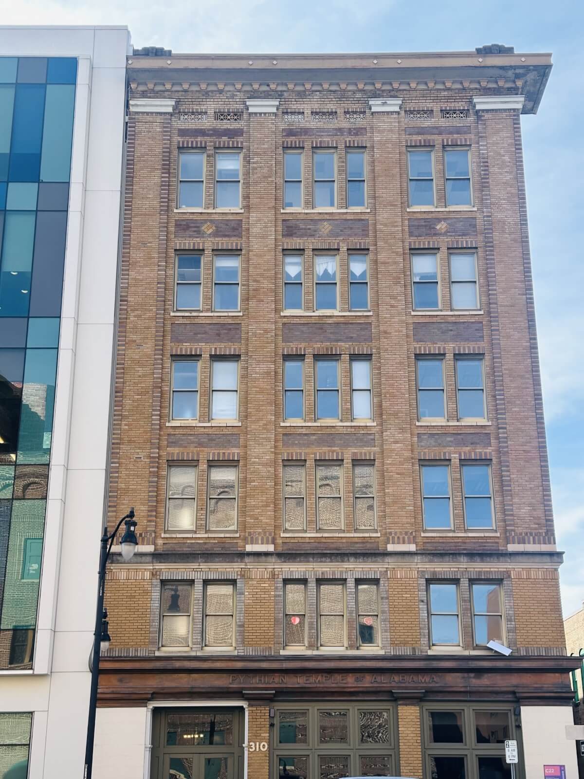 The historic six-story brick building features a decorative cornice and stands bordered by modern white and glass structures. A streetlamp is visible in front.