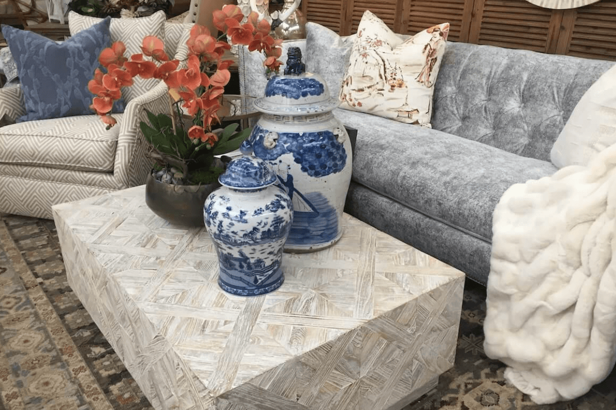 A living room with a patterned coffee table—sourced from one of the best furniture and antique stores in Nashville—holds blue and white ceramic jars and an orange orchid, set before a gray couch with cushions and a white throw.