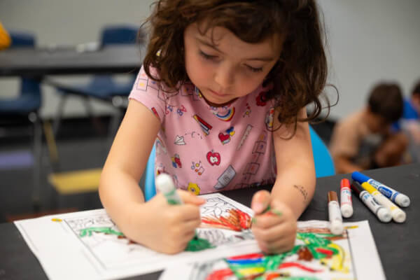 A young child with curly hair, in a pink shirt, colors on a drawing with markers at a table. Other supplies and children are visible in the background.