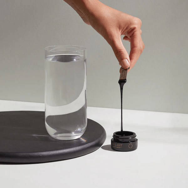 A hand carefully dips a stick into a jar of black substance, positioned next to a glass of clear water on a circular mat.