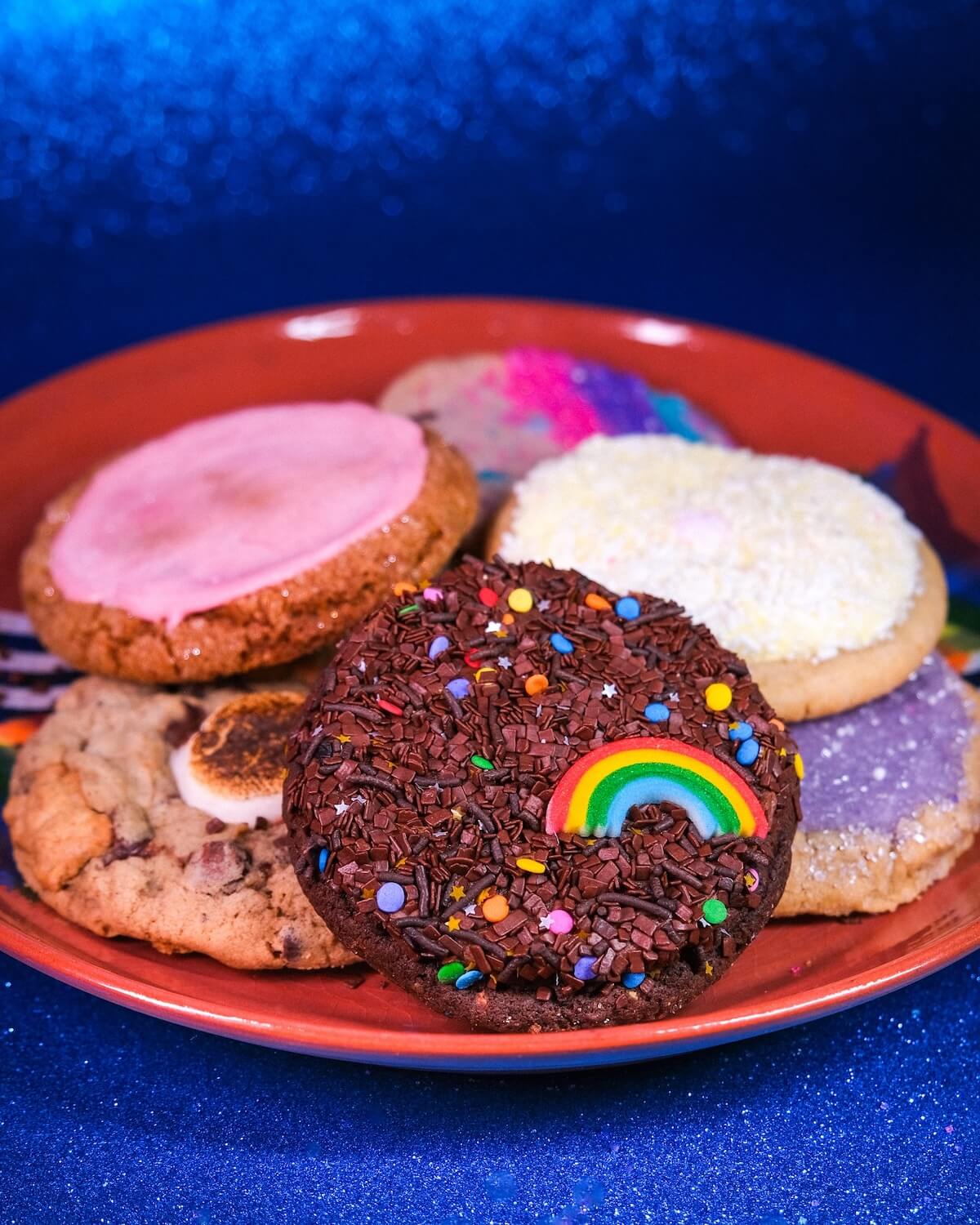 A plate with six assorted decorated cookies, including one with chocolate sprinkles and a rainbow candy, makes the SB Hot List for January 2025 against a vibrant blue background.