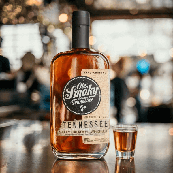 A bottle of Ole Smoky Tennessee Salty Caramel Whiskey graces the SB Hot List this January 2025. It stands alongside a small glass on a reflective surface, set against a backdrop of blurred lights in an indoor setting.