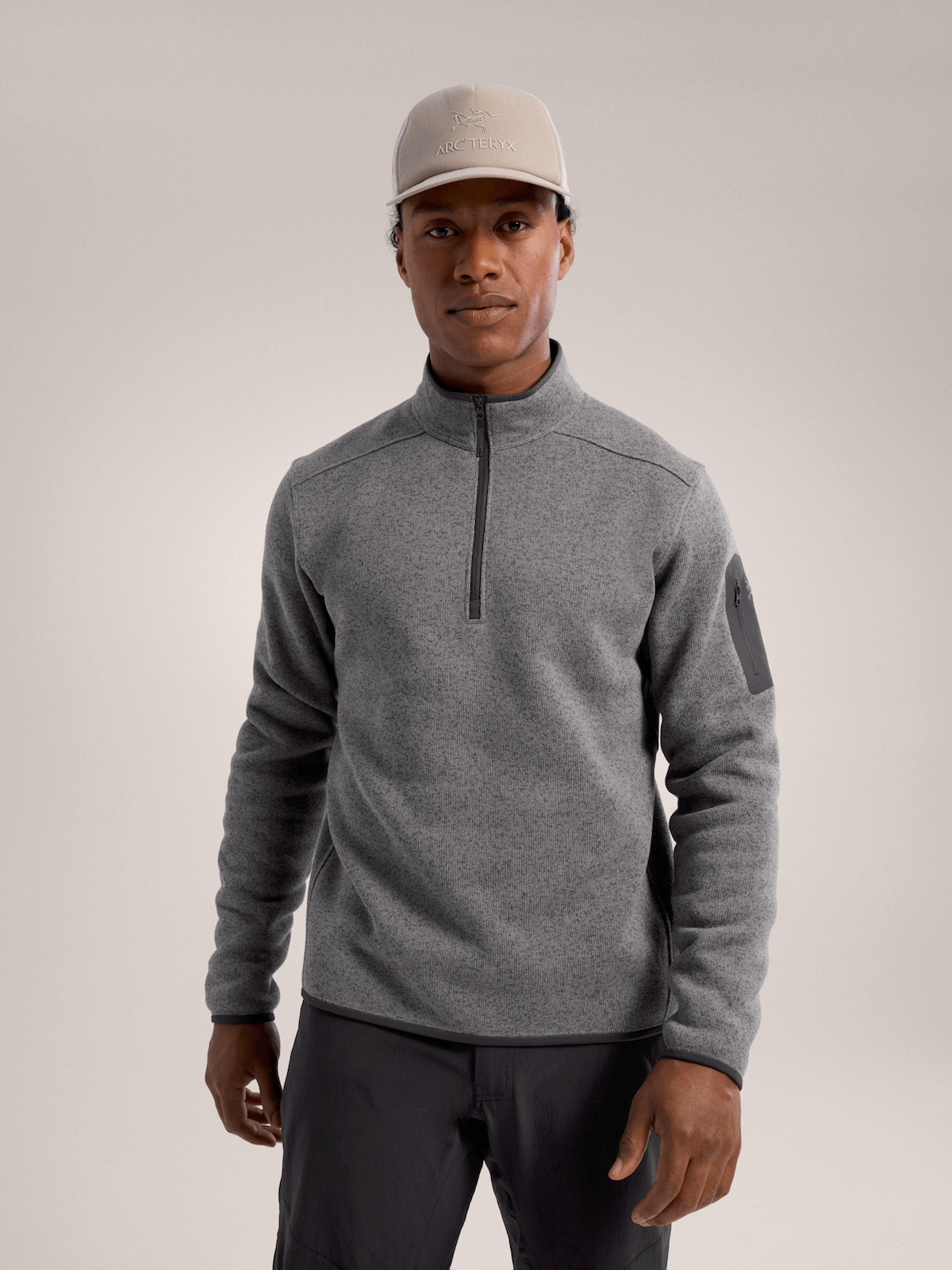 Person wearing a gray half-zip fleece jacket, gray cap, and dark pants against a plain background.