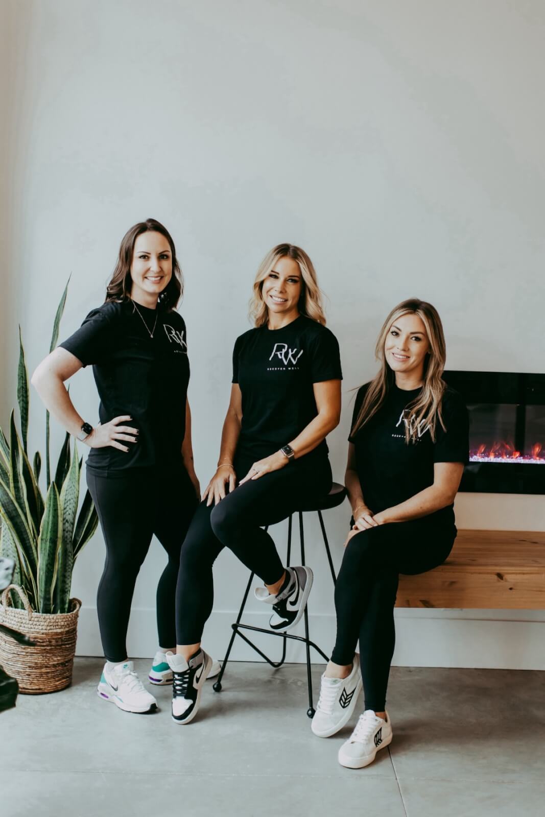 Three women in black t-shirts and leggings pose by an indoor fireplace, embodying the spirit of Recover Well. One sits on a stool, while the others stand gracefully. A lush potted plant adds a touch of nature to the scene.