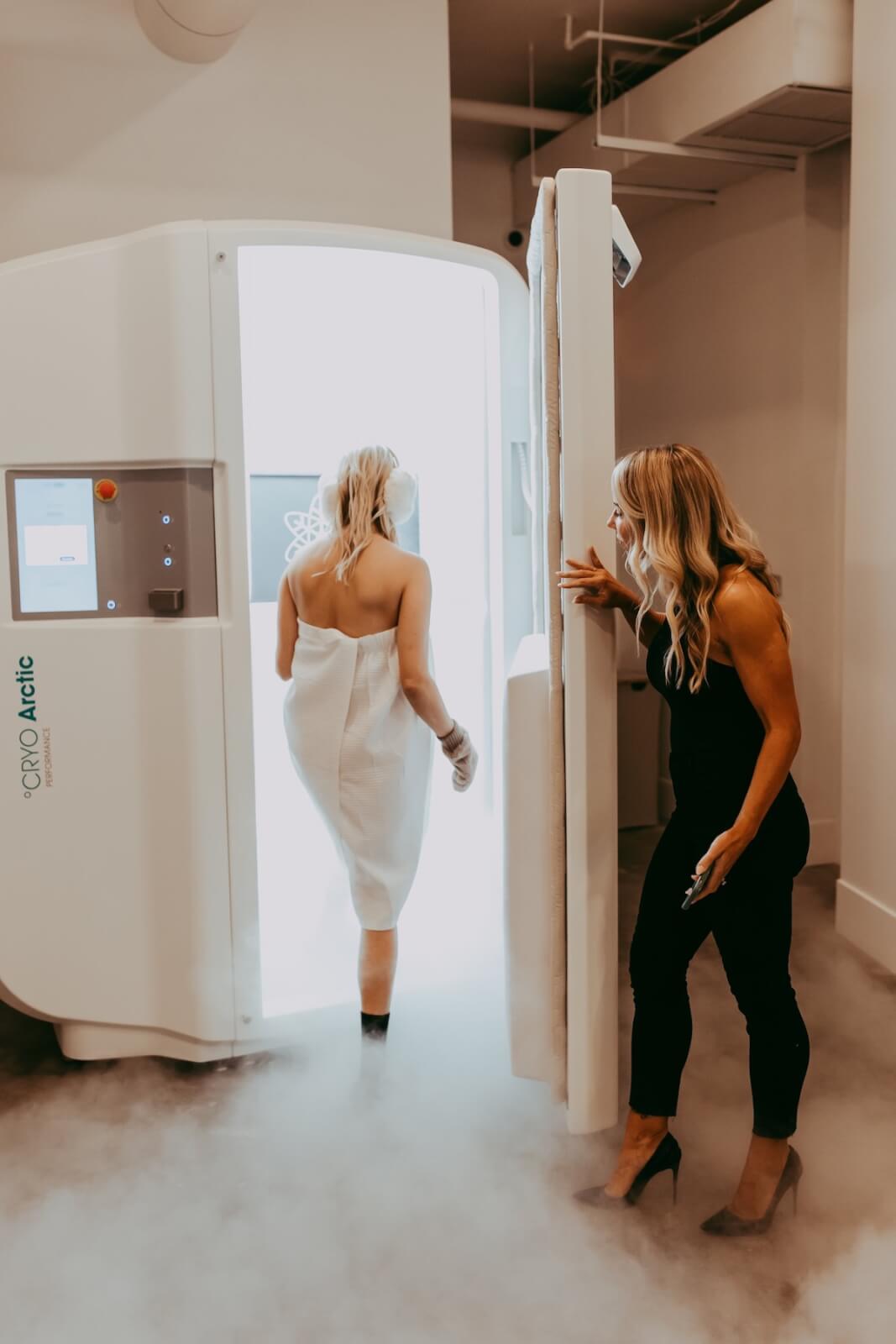 A woman wearing a towel enters a cryotherapy chamber, embraced by cool mist, while another woman observes.
