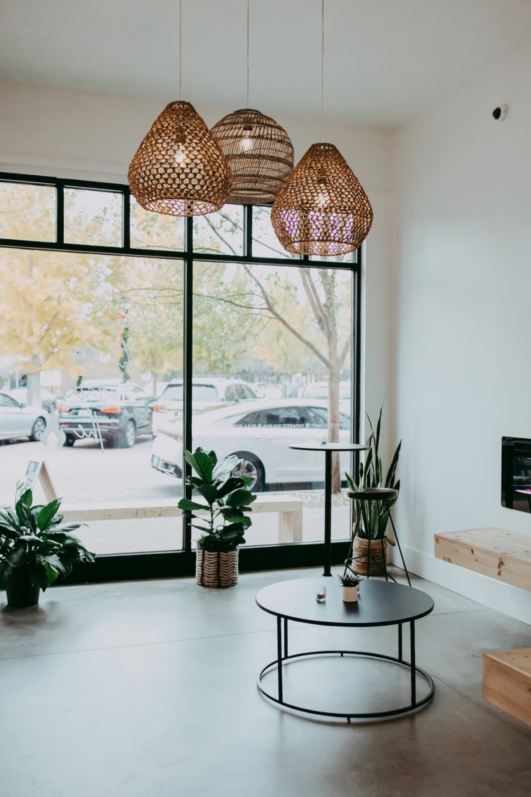 This minimalist room in Louisville features wicker pendant lights, a round coffee table, and potted plants. Large windows overlook parked cars and trees outside, while an infrared sauna adds a touch of wellness to the serene space.