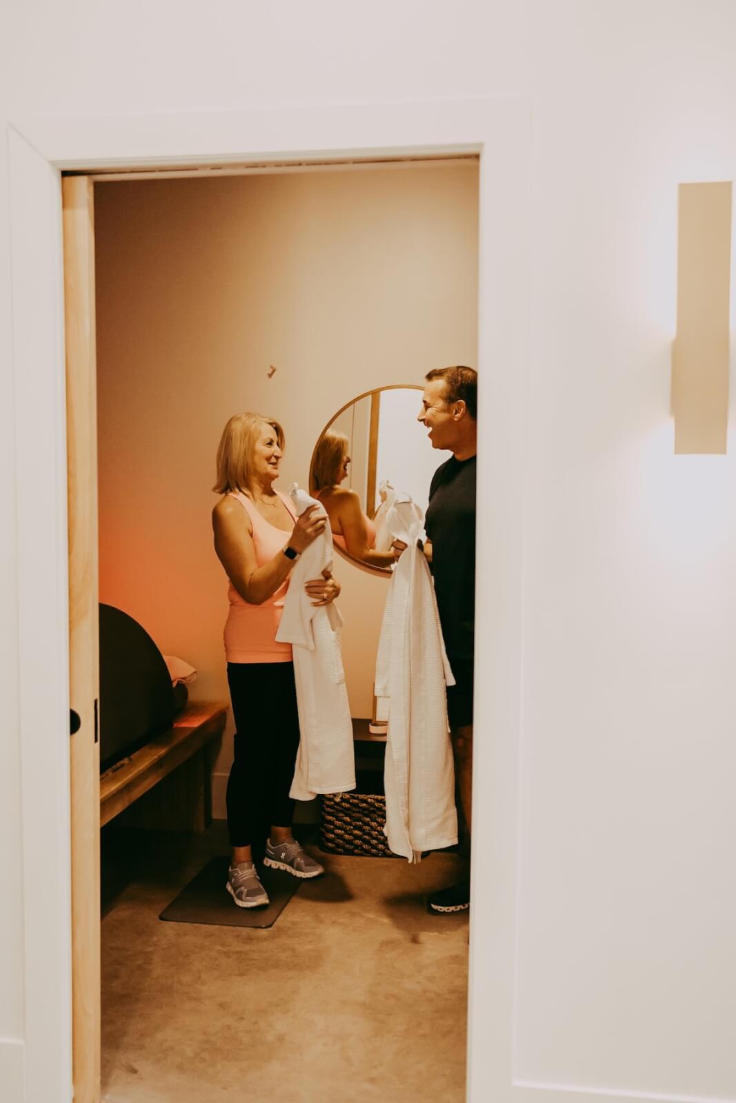 A man and a woman stand in a room with a mirror, holding white towels, engaging in conversation about their recent cryotherapy session.