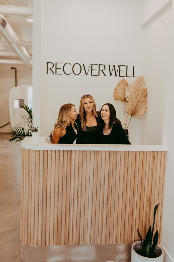 Three people stand behind a wooden reception desk under a sign that reads "RECOVER WELL." They are smiling and dressed in black, ready to introduce visitors to the rejuvenating benefits of cryotherapy and infrared sauna treatments.