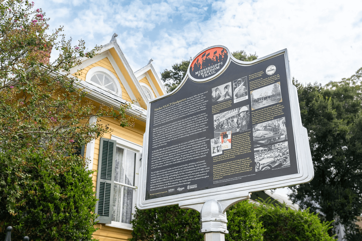 A historical marker stands before a yellow house with green shutters, offering insights into a significant event or figure along Mississippi's Freedom Trail. Surrounded by trees, it sits under a partly cloudy sky, guiding visitors through the echoes of history.
