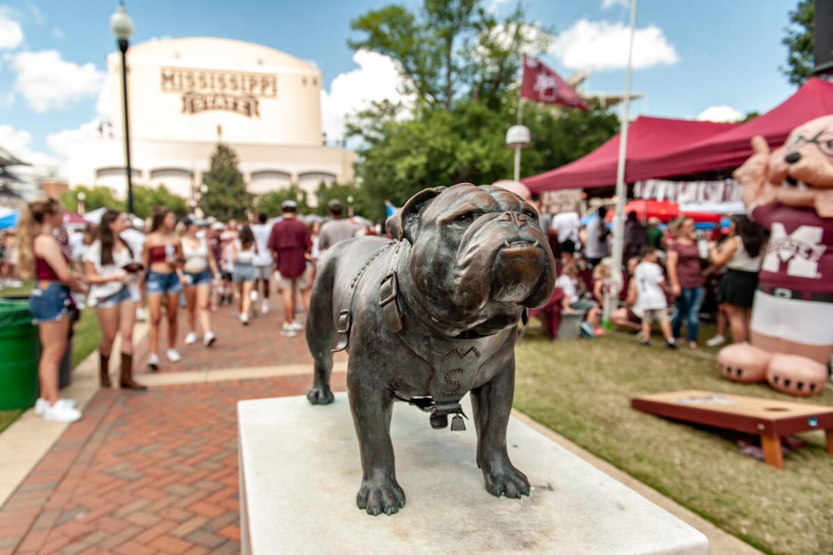 A bronze bulldog statue stands proudly in front of Mississippi State University, surrounded by people and tents on a sunny day, capturing the lively spirit of this historic Mississippi landmark.
