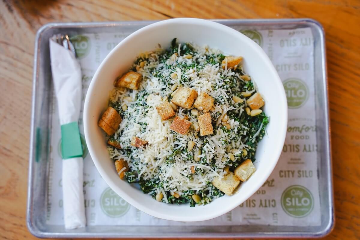 A white bowl of kale salad topped with grated cheese, croutons, and seeds, served on a tray with wrapped utensils on the side—one of the best salads in Memphis.