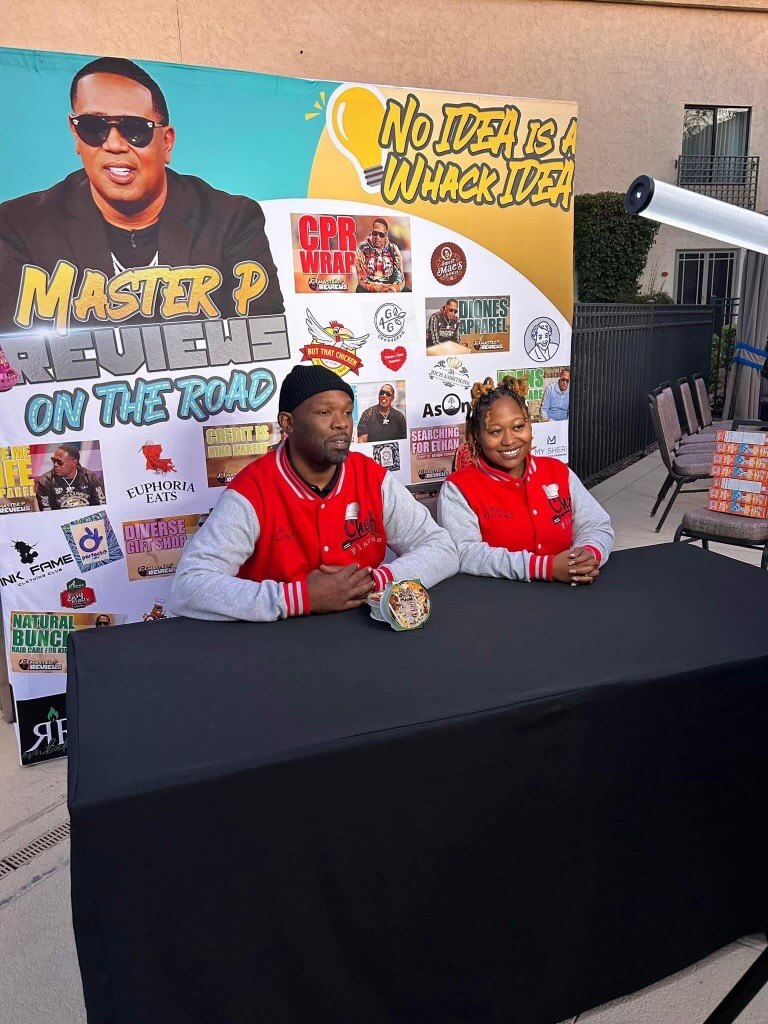 Two people in red jackets sit at a table with various products on display. Behind them, a large promotional banner proudly showcases Chef Flavas' latest culinary creations, as Regina Moore discusses her Memphis roots and cooking inspirations.