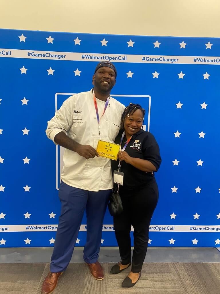 Two people stand smiling in front of a blue backdrop with white stars and "#WalmartOpenCall." Regina Moore holds a yellow envelope with "Got a deal!" printed on it.