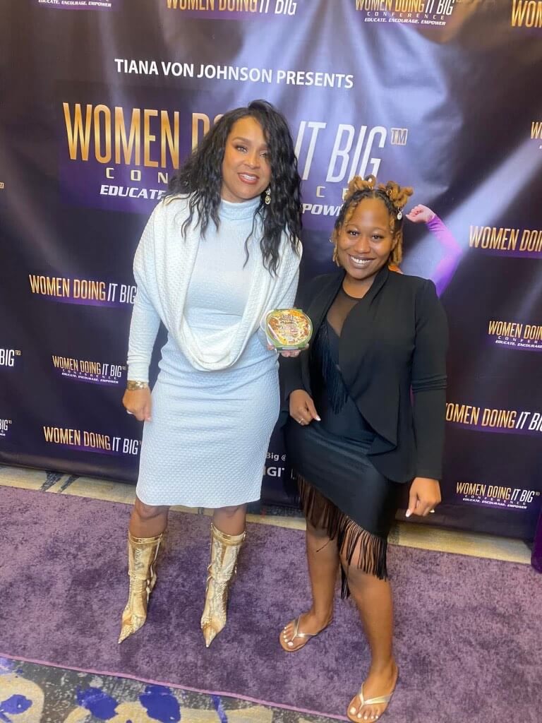 Two women pose at the "Women Doing It Big" conference in Memphis, with one proudly holding an award. They stand in front of a banner showcasing the event’s name, embodying the spirit of achievements celebrated by remarkable individuals like Regina Moore.