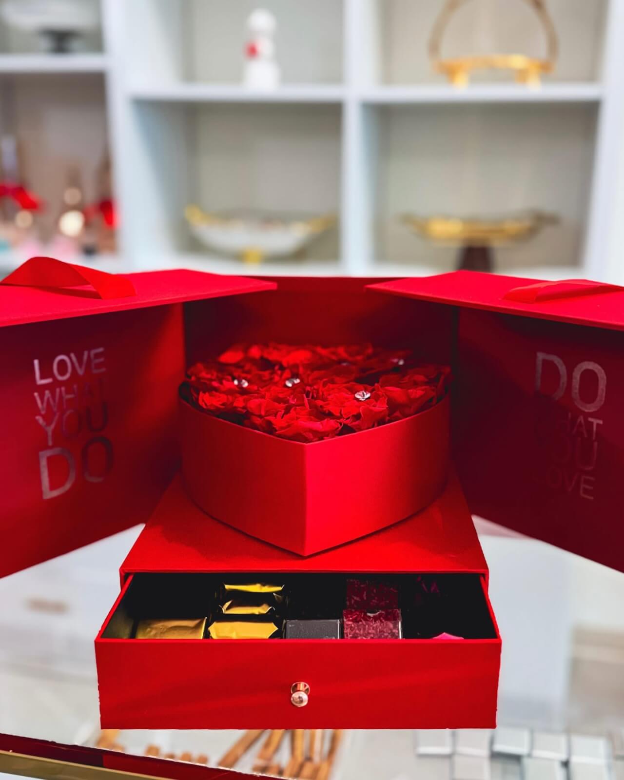 The red box marked "LOVE WHAT YOU DO" opens to reveal a heart-shaped arrangement of red roses and a drawer filled with assorted chocolates. Perfect for February discoveries, this exquisite gift set brings warmth against a blurred background of your cherished finds.