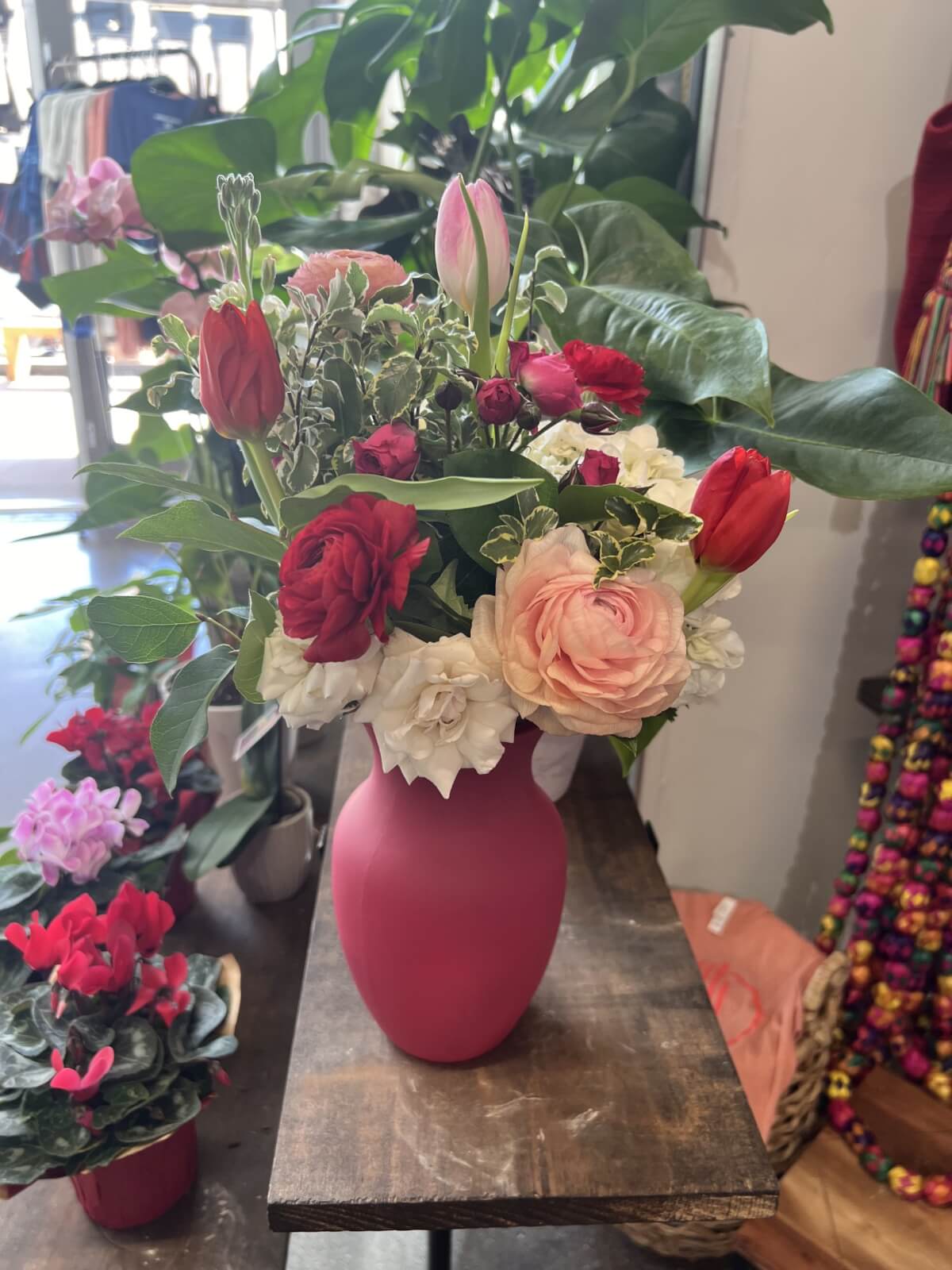In a charming shop setting reminiscent of Memphis in February, a vase brimming with red and pink flowers, including roses and tulips, graces a wooden table, surrounded by more exquisite plant finds.