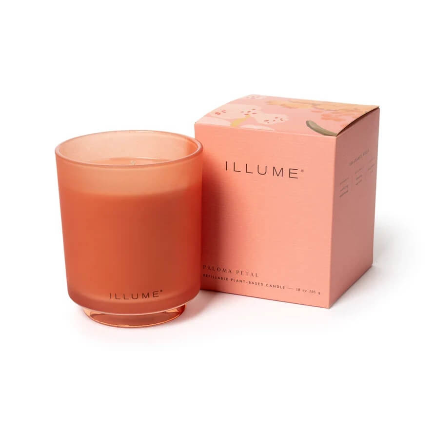 An orange candle in a glass jar sits elegantly beside a matching orange cardboard box labeled "ILLUME" and "Paloma Petal," reminiscent of February blossoms. Like a hidden gem in Memphis, the set invites warmth and discovery with its unique allure.