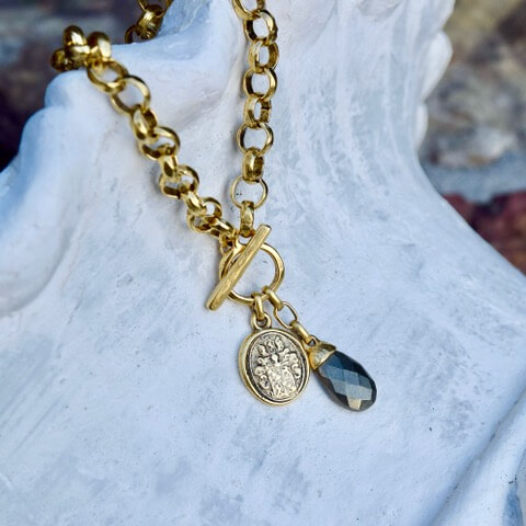 The Gold chain necklace, adorned with a toggle clasp, features a gold medallion and faceted teardrop pendant. This stunning piece is beautifully displayed on a textured white surface.