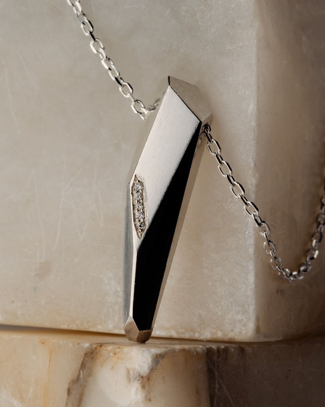 A silver geometric pendant with a small diamond detail, suspended on a silver chain, radiates against a marble background.