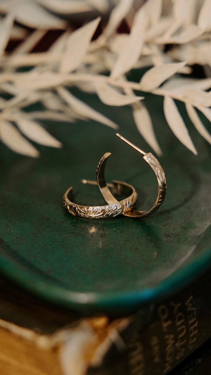 Two silver hoop earrings with engraved patterns rest on a green surface, accompanied by dried white leaves. This elegant display from Memphis Jewelry Brands captures the essence of style and sophistication.
