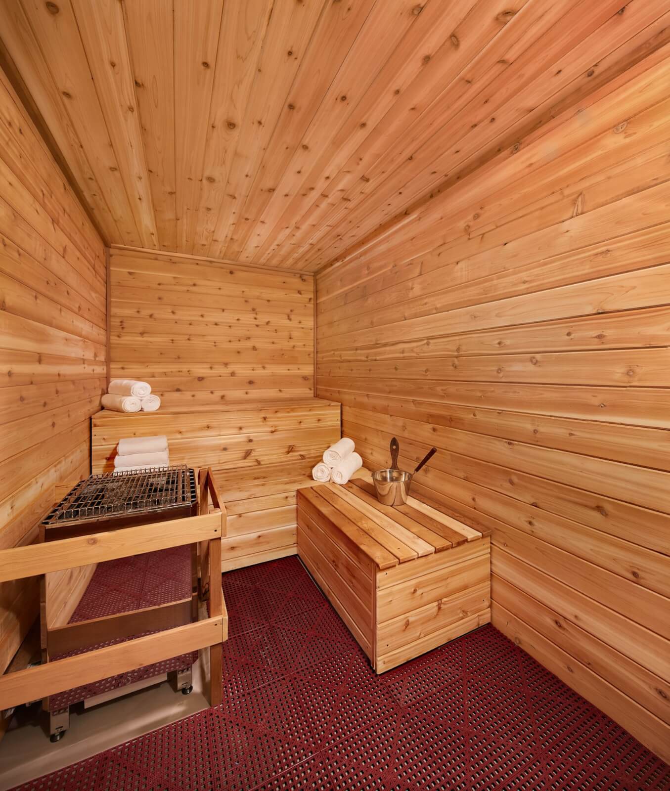Memphis-style wooden sauna interior with folded white towels on a shelf and bench. A metal bucket with a ladle rests on another wooden surface, while the floor is covered with vibrant red grid-like mats.