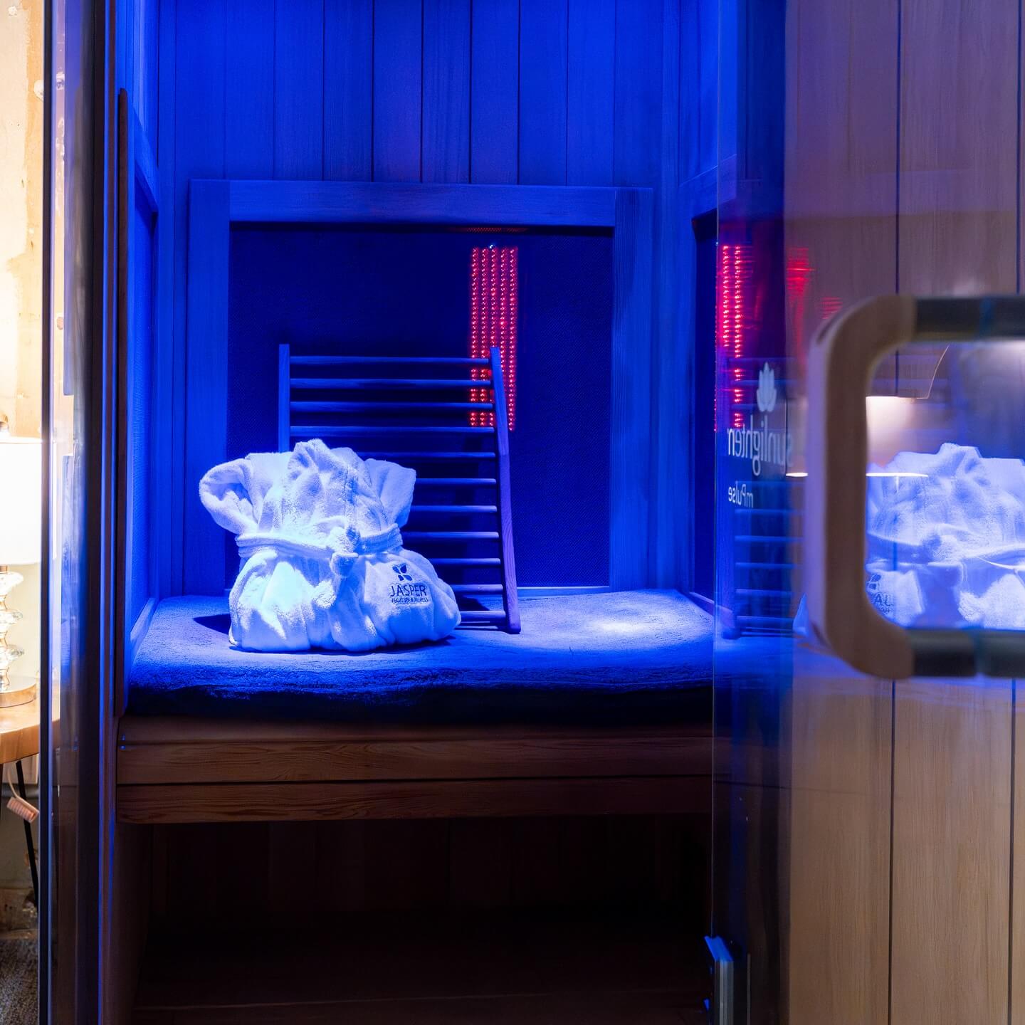 Memphis-inspired blue-lit sauna room with a wooden bench, folded white robe, and a towel on top. A glass door is visible on the right.