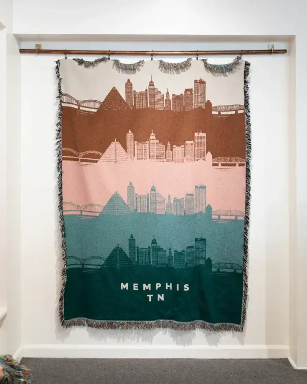 A woven tapestry elegantly captures the Memphis, TN skyline in brown, pink, and teal hues. Displaying iconic architectural landmarks with fringe edges, this stunning piece is hung on a rod in a white-walled room. Discover January 2025's finest artistic expression with Memphis FINDS.