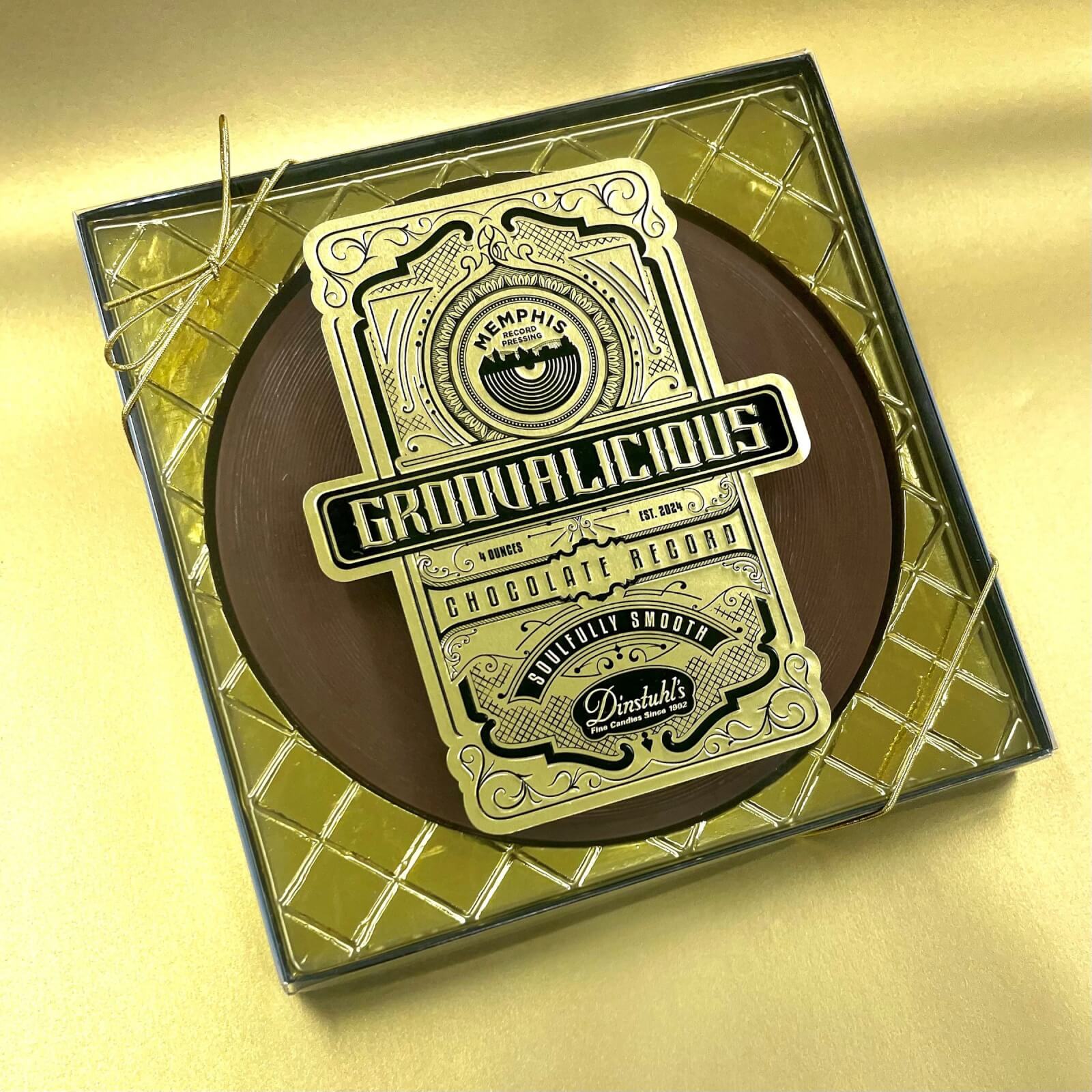 Square box with a chocolate shaped like a vinyl record inside. Label reads "Groovalicious Chocolate Record," with decorative gold and black design on a gold background.