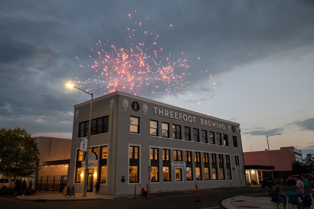 Fireworks burst above a building labeled "Threefoot Brewing" during twilight, with the sounds of Meridian music filling the air as people gather below on the street.
