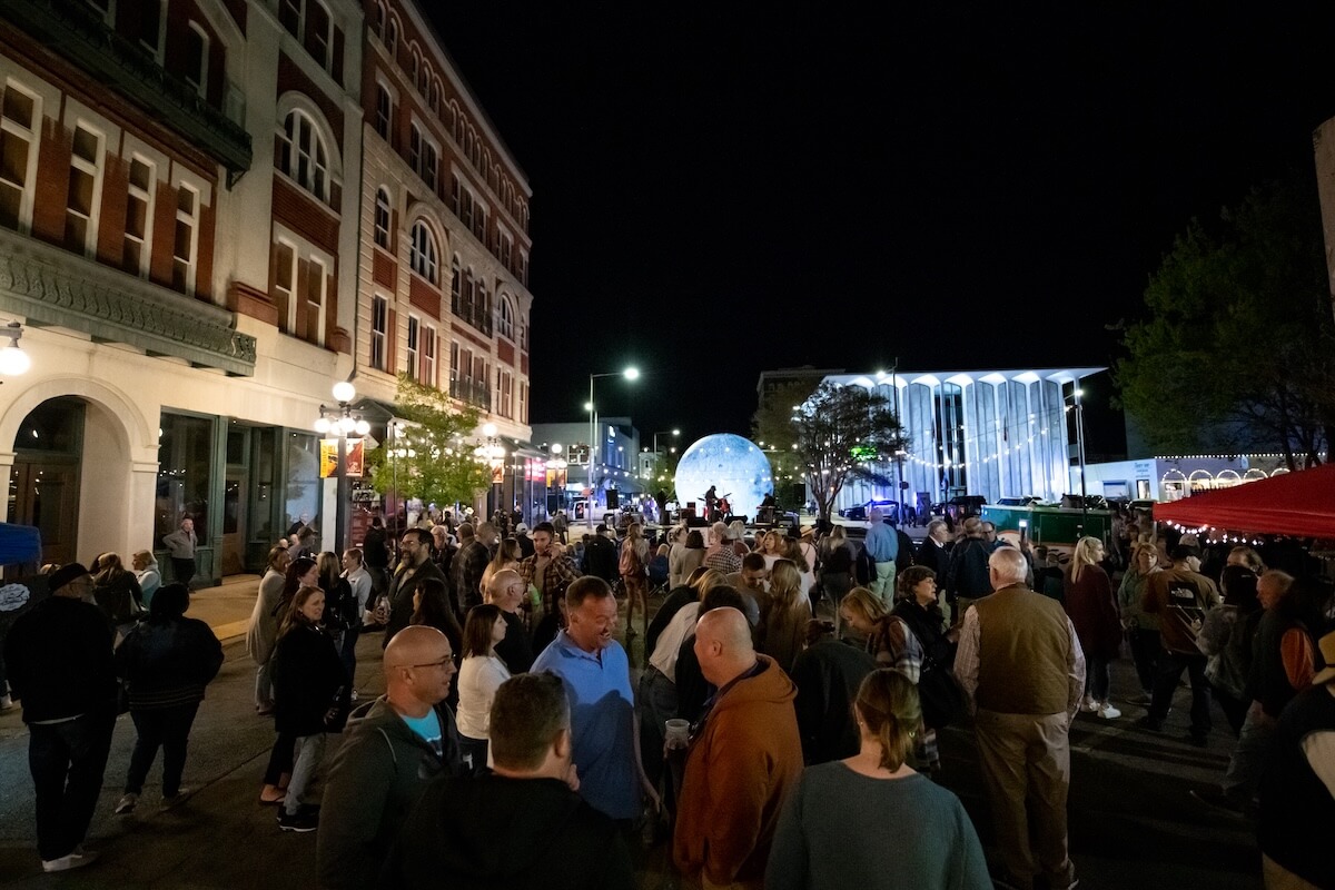 A large crowd gathers at a nighttime outdoor street event, the air filled with meridian music. In the backdrop, buildings stand tall, while a lit moon glows against the dark sky.