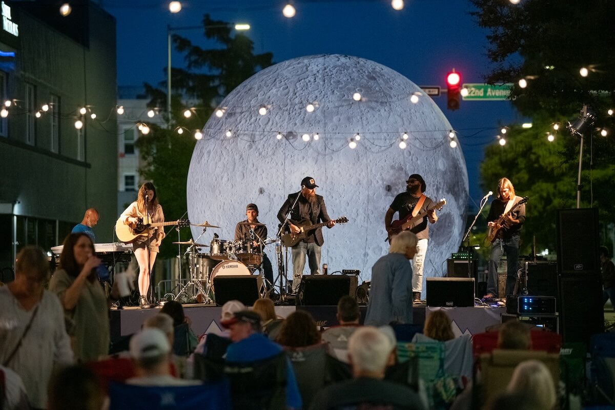 A band performs on an outdoor stage with a large moon backdrop, captivating the audience with their meridian music. Spectators are seated in the foreground beneath twinkling string lights.