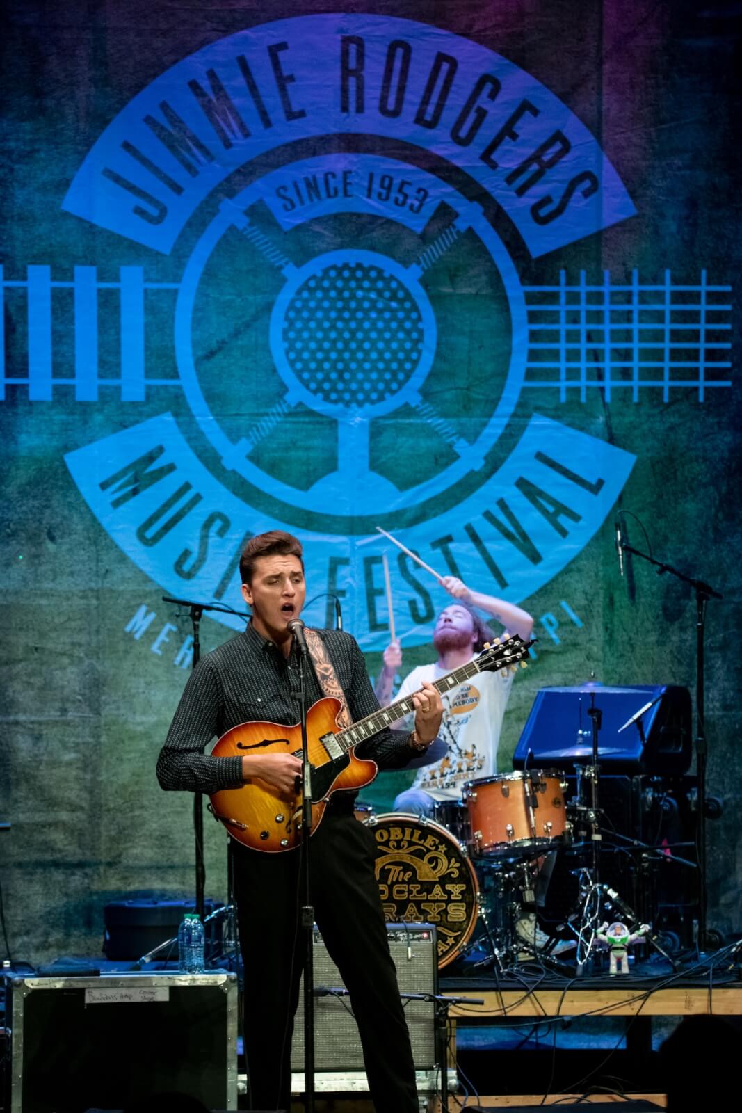 A musician plays electric guitar on stage at the Jimmie Rodgers Music Festival, injecting a vibrant energy into Meridian's music scene, with a drummer performing in the background.