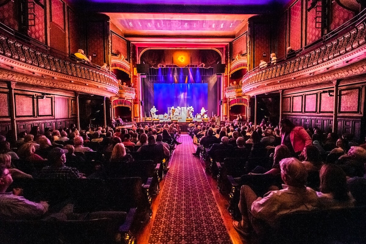 An audience in a theater watches a performance on stage, enveloped by the mesmerizing tones of meridian music. The theater features ornate balconies and vibrant lighting, creating an enchanting atmosphere.