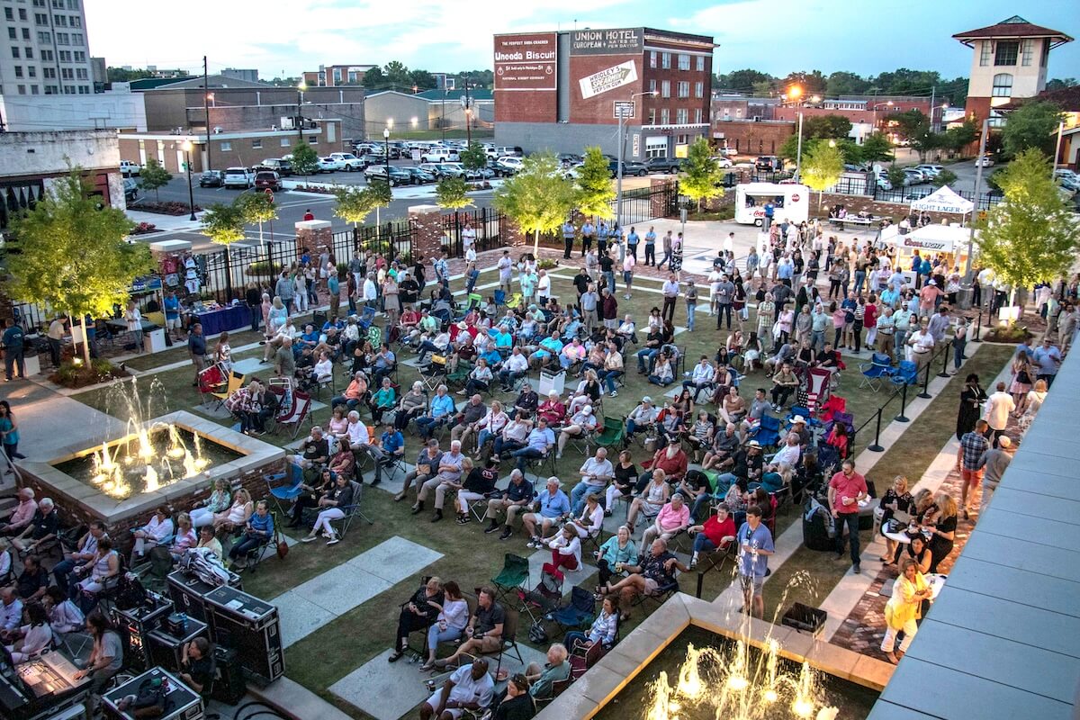 A large crowd gathers at Sipp & Savor, surrounded by buildings and trees, while the aroma from nearby food stands wafts through the air.
