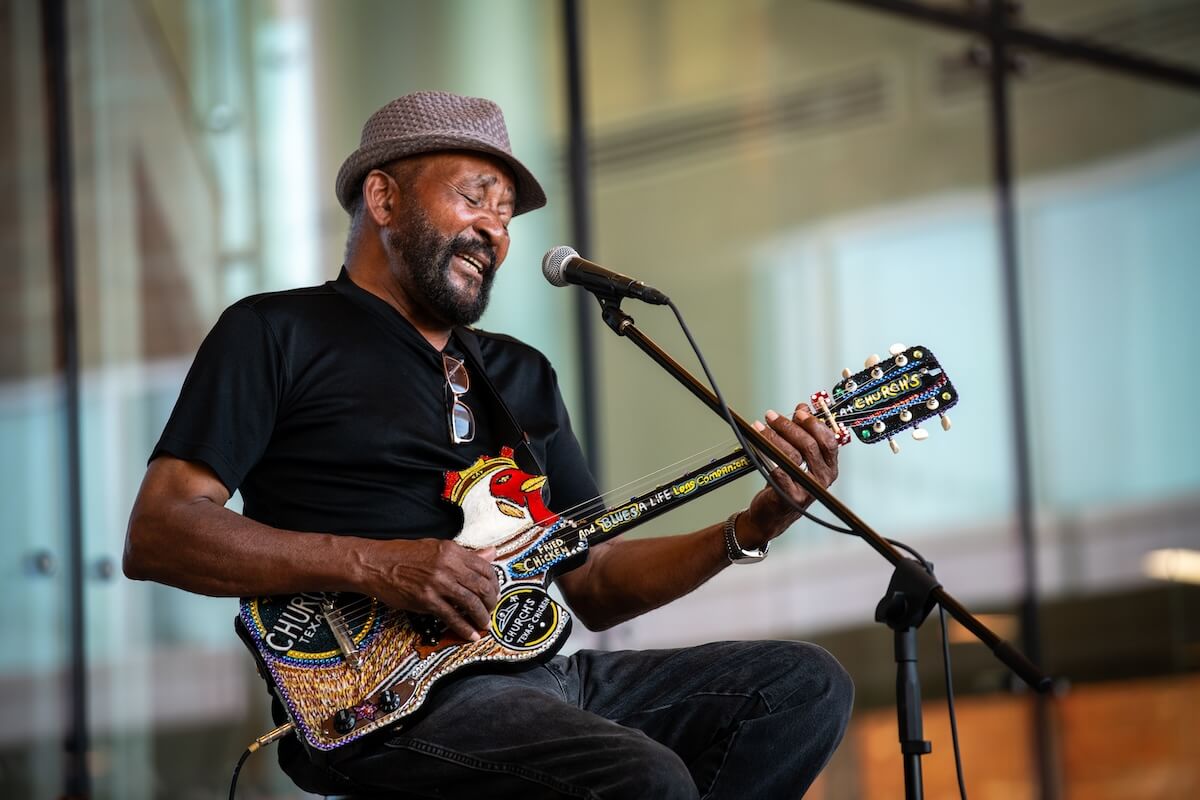 A man wearing a hat and sunglasses strums an elaborately decorated guitar while singing meridian music into a microphone.