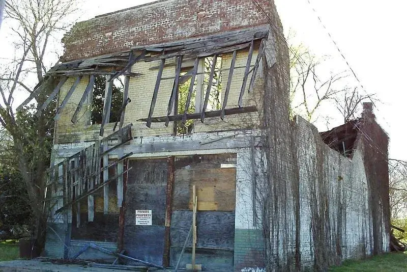 The dilapidated brick building, with its partly collapsed roof and boarded-up windows, stands hidden behind overgrown vegetation. A "Private Property" sign guards its history.